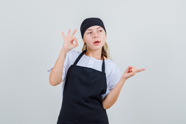 Young waitress showing ok sign and pointing to side in uniform and apron