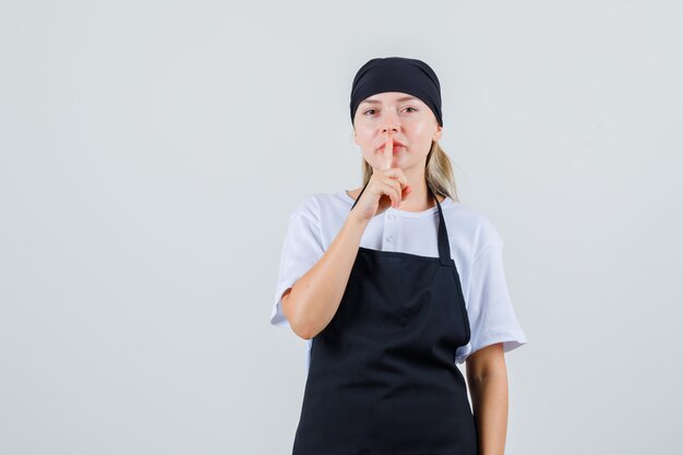 Молодая официантка показывает жест молчания в униформе и фартуке и смотрит осторожно