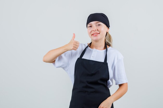 Молодая официантка показывает палец вверх в униформе и фартуке и выглядит весело