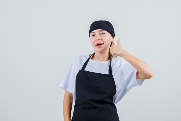 Young waitress showing thumb up in uniform and apron and looking pleased