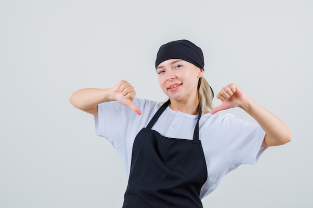 Free photo young waitress showing thumbs down in uniform and apron and looking cheery