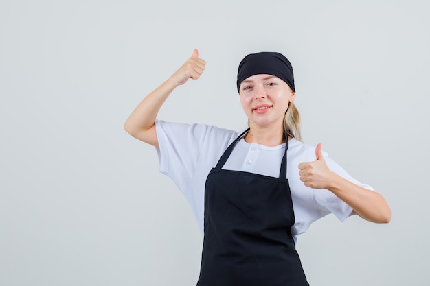 Young waitress showing thumbs up in uniform and apron and looking cheerful