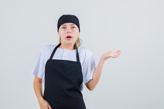 Young waitress spreading raised palm aside in uniform and apron and looking puzzled