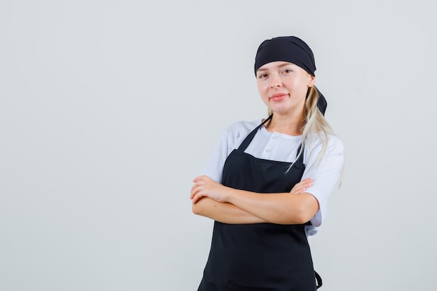 Giovane cameriera in piedi con le braccia incrociate in uniforme e grembiule e guardando allegra