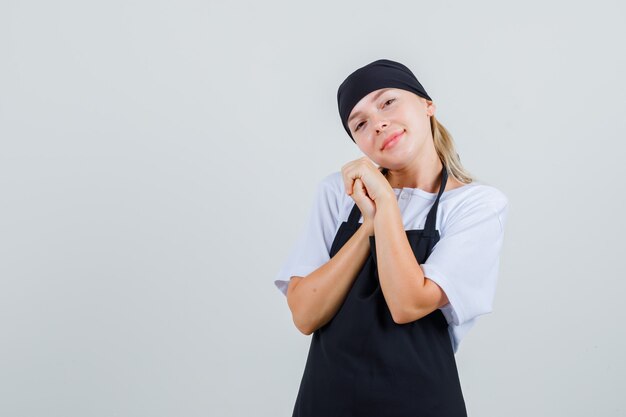 Young waitress in uniform and apron bowing head on clasped hands and looking beloved