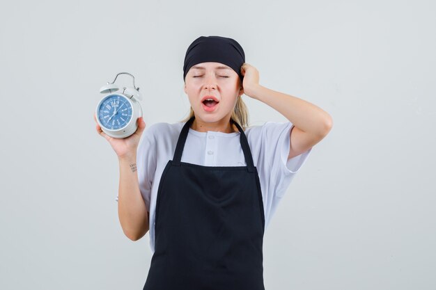 Free photo young waitress in uniform and apron holding alarm clock and looking forgetful