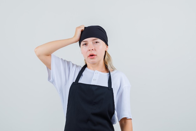 Free photo young waitress in uniform and apron holding hand on head and looking pensive