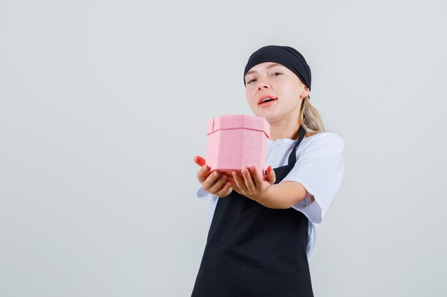 Young waitress in uniform and apron holding present box and looking optimistic