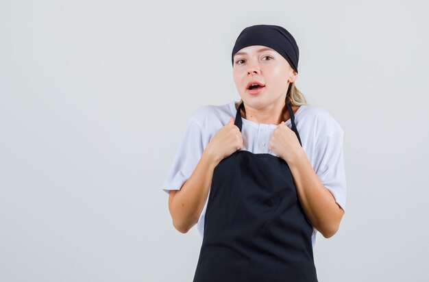 Young waitress in uniform and apron keeping fists on chest and looking amazed
