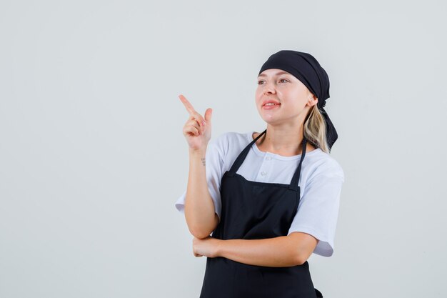 Young waitress in uniform and apron pointing to side and looking cheerful