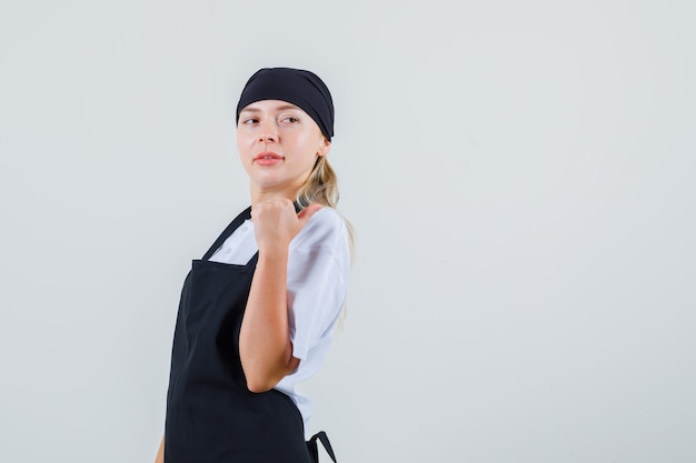 Young waitress in uniform and apron pointing thumb back and looking confident