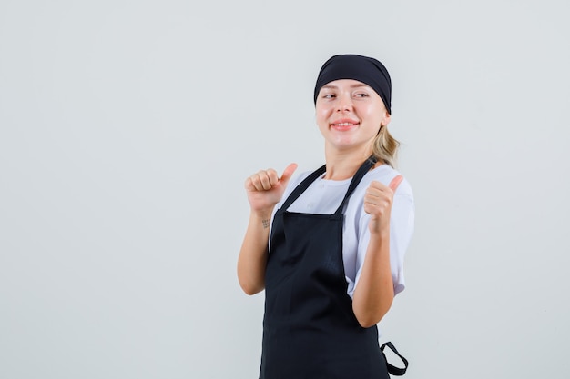 Young waitress in uniform and apron pointing thumbs back and looking cheerful