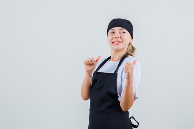Молодая официантка в униформе и фартуке, указывая большими пальцами назад и выглядит веселой