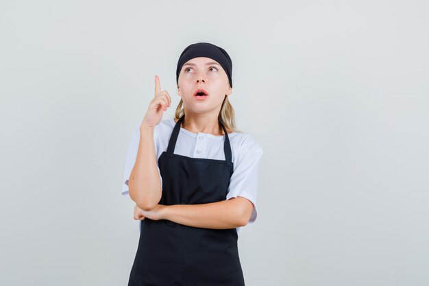 Young waitress in uniform and apron pointing up and looking curious
