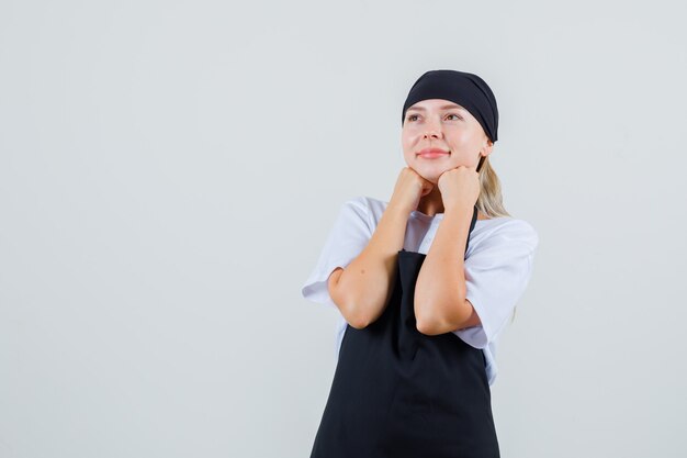 Giovane cameriera in uniforme e grembiule che tiene i pugni sul mento e sembra speranzosa