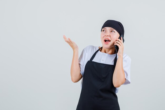 携帯電話で話し、驚いて見える制服とエプロンの若いウェイトレス