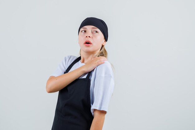 Young waitress in uniform and apron touching shoulder while looking back