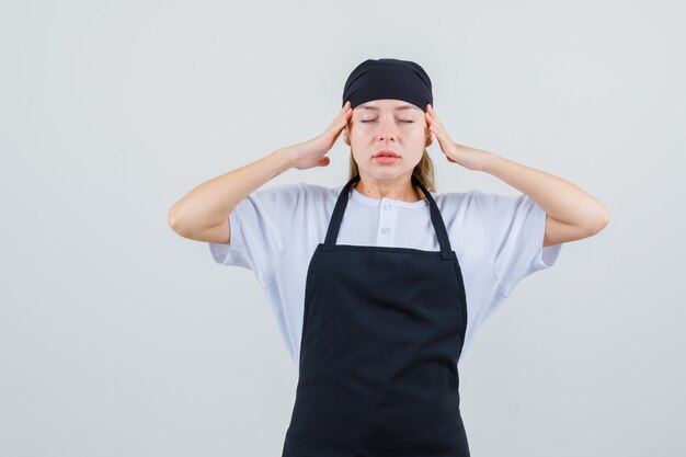 Young waitress in uniform and apron touching temples with fingers and looking tired