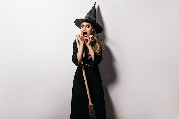 Young witch in hat posing in halloween with scary face expression. Indoor photo of shocked blonde female model in wizard costume.
