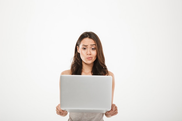 young woman 30s looking on screen of her silver notebook thinking or expressing misunderstanding with face, isolated over white wall