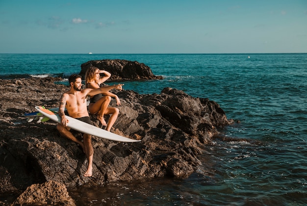 무료 사진 바다 근처 바위에 서핑 보드와 함께 측면을 가리키는 젊은 여자와 남자