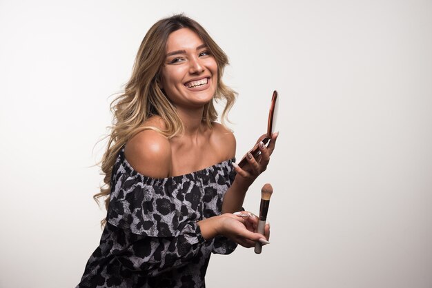 Young woman applying bronzer on white wall.