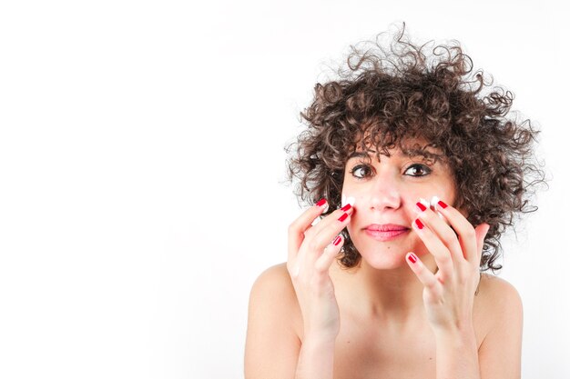 Young woman applying cosmetic cream on her face against white background