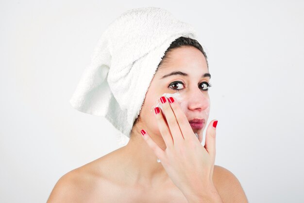 Young woman applying cream on face with white towel over her head