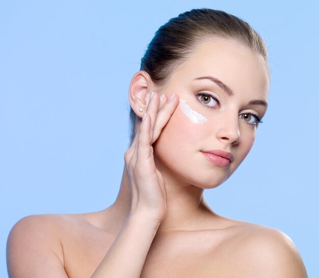 Young woman applying cream on her face on blue