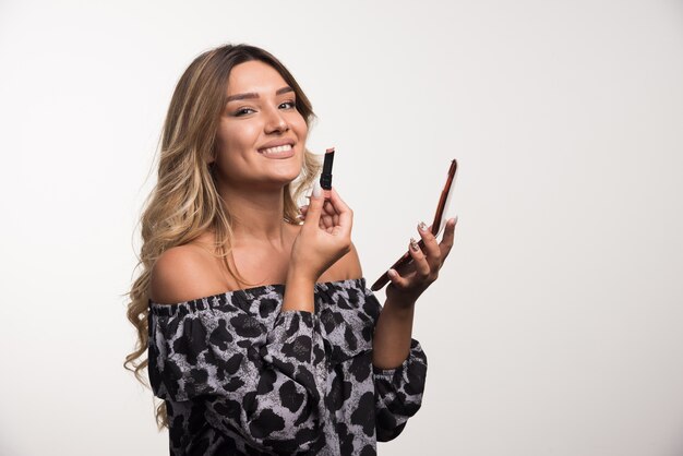 Young woman applying lipstick on white wall.