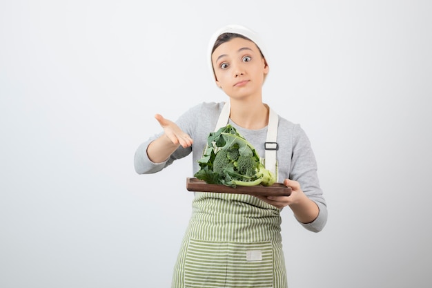 Giovane donna in grembiule che tiene piatto di broccoli su bianco