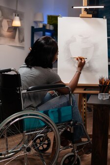スケッチ​グラフィック​鉛筆​を​使用して​キャンバス​に​花瓶​を​描く​障害​を​持つ​若い​女性​アーティスト​。​自宅​の​アトリエ​で​アート​レッスン​の​ため​の​絵画​ツール​を​使用して​イラスト​を​描く​で​働いている​車椅子​の​学生