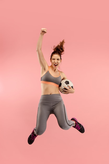 La giovane donna come calciatore di calcio che salta e calcia il pallone in studio su uno sfondo rosso.