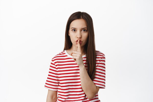 Young woman asking to be quiet, keep secret, hushing with finger on lips and frowning, stay silent, taboo sign, standing against white wall.