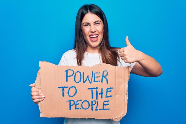 Young woman asking for social movement holding banner with power to the people message smiling happy and positive thumb up doing excellent and approval sign