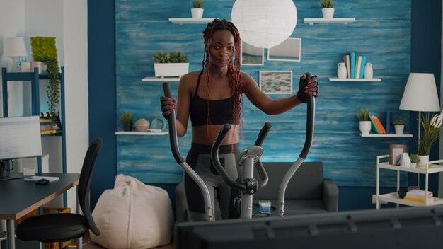 Young woman athletic training on elliptical bike practicing cardio sport watching aerobic video on tv during bodyweight morning routine in living room