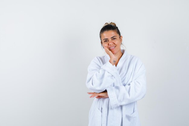 Young woman in bathrobe holding hand on cheek and looking pretty