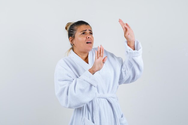 Young woman in bathrobe keeping hands in defending pose and looking scared
