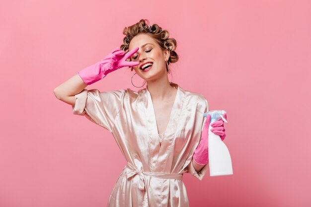 Young woman in bathrobe and rubber gloves shows peace sign and holds detergent