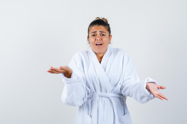 Young woman in bathrobe showing helpless gesture and looking confused