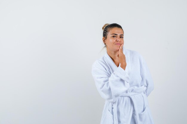 Young woman in bathrobe touching chin with finger and looking cute