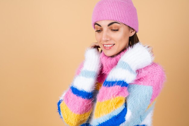 Young woman on beige in a bright multi-colored cozy knitted sweater and a hat comfortably hugs herself, warmth and comfort