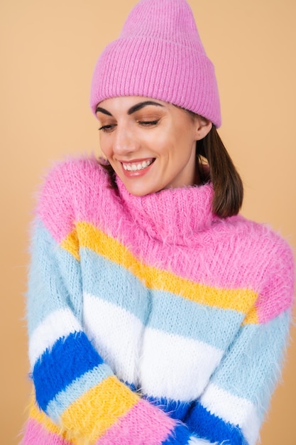 Young woman on beige in a bright multi-colored cozy knitted sweater and a hat comfortably hugs herself, warmth and comfort