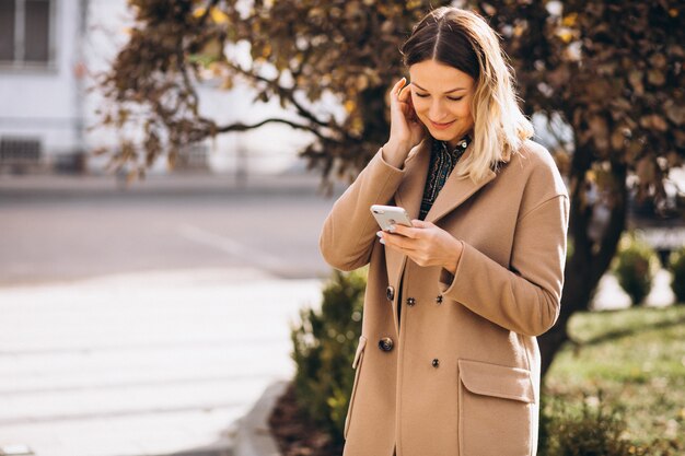 通りの外の電話を使用してベージュのコートの若い女性