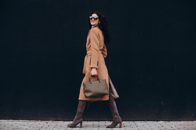 Young woman in beige coat walking outdoors on black wall background