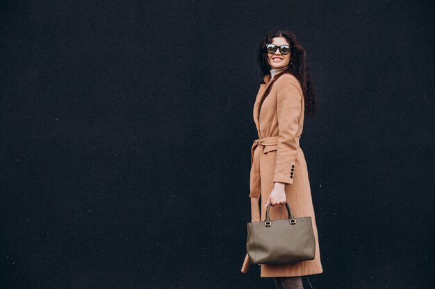 young woman in beige coat walking outdoors on black wall background