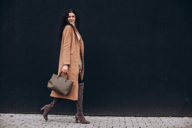 Young woman in beige coat walking outdoors on black wall background