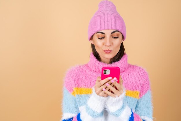 Young woman on beige in a knitted sweater and a hat cute thoughtfully typing a message on the phone, biting her lip
