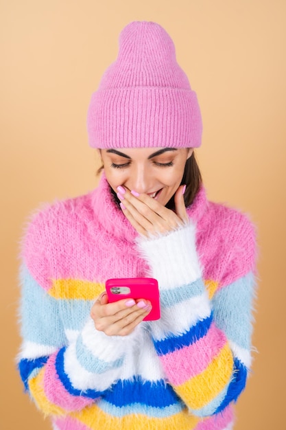 Young woman on beige in a knitted sweater and a hat with a mobile phone laughing with surprise and fun, reading messages, jokes, congratulations