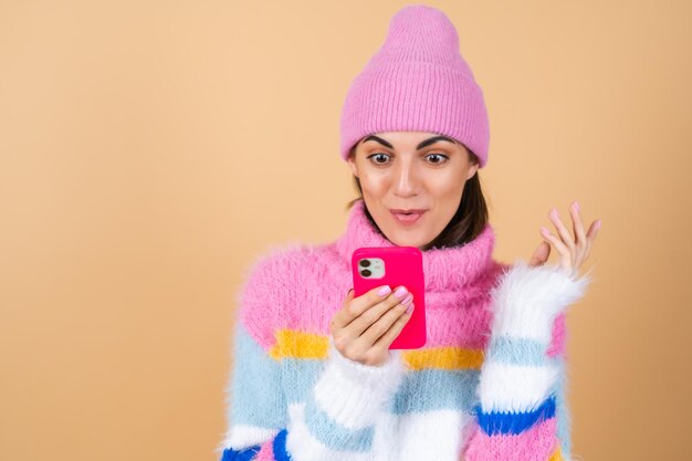 Young woman on beige in a knitted sweater and a hat with a mobile phone laughing with surprise and fun, reading messages, jokes, congratulations